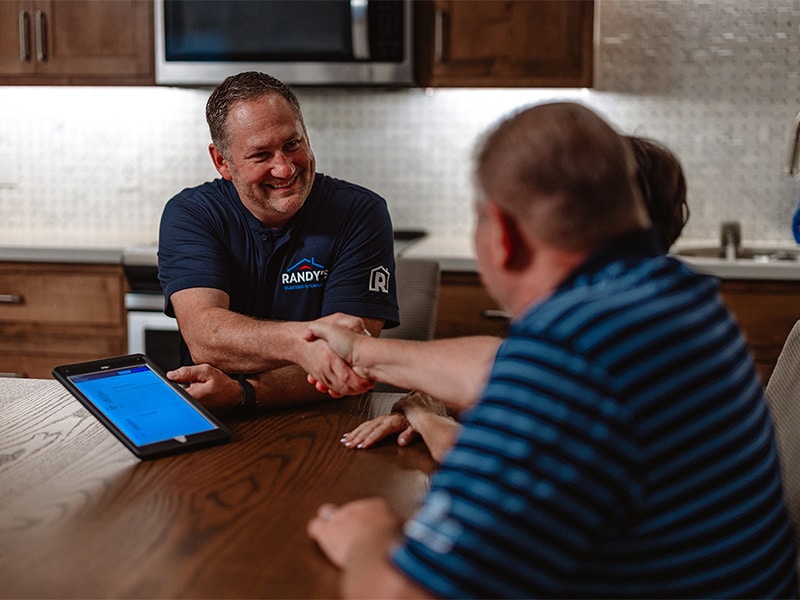 Randy shaking hands with a couple over a table with a tablet, inside a modern kitchen setting.