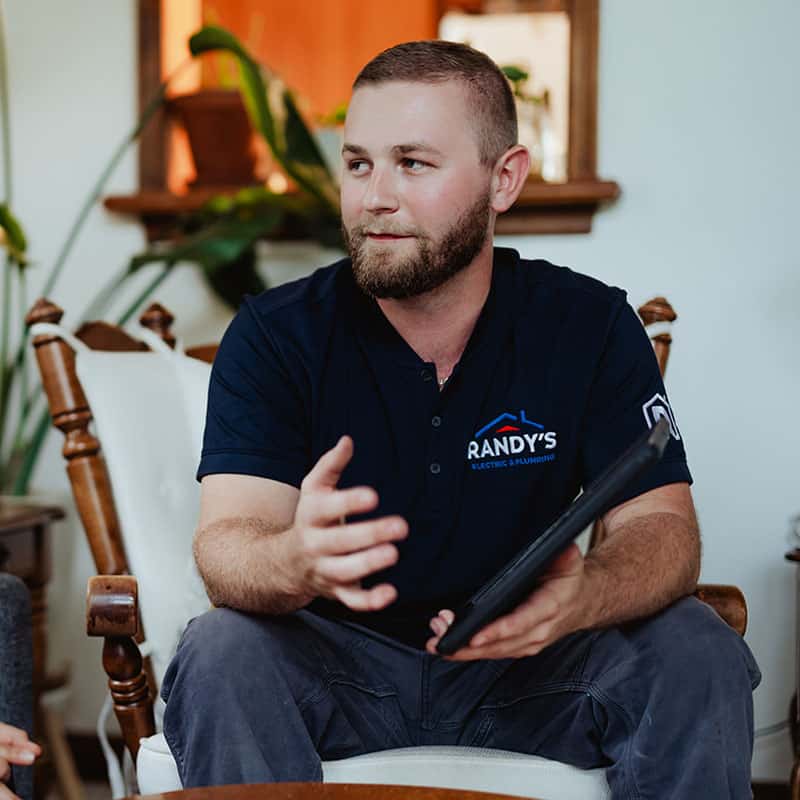 A man in a navy uniform from Randy's Electric & Plumbing, holding a tablet, gestures while seated indoors.